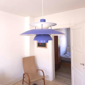 May include: A multi-tiered pendant light fixture featuring a series of stacked, circular shades in a vibrant purple hue. The design includes a white top shade and a central white interior component, all suspended from a single white cord.