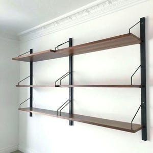 Puede incluir: Estantería de pared de tres niveles con estantes de madera oscura y soportes de metal negro. La estantería está fijada a una pared blanca. Cada estante tiene unos 25 cm de profundidad.