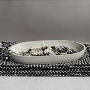 Könnte beinhalten: Eine hellgraue ovale Schale aus Beton mit einer Perlen- und Silberkettenkette und einem Silberkettenarmband darin. Die Schale liegt auf einem schwarz-weißen gewebten Stoff.