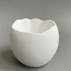 Op de afbeelding: Een witte keramische plantenpot met een golvende, onregelmatige rand. De pot heeft de vorm van een ei en heeft een gladde, matte afwerking.