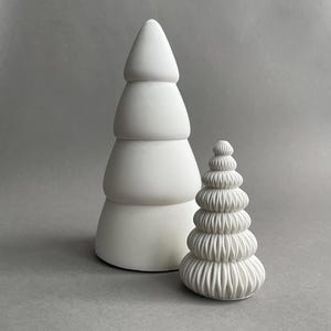 Puede incluir: Dos &aacute;rboles de Navidad de cer&aacute;mica blanca. El &aacute;rbol m&aacute;s grande tiene un dise&ntilde;o liso y redondeado. El &aacute;rbol m&aacute;s peque&ntilde;o tiene un dise&ntilde;o texturizado y con volantes.