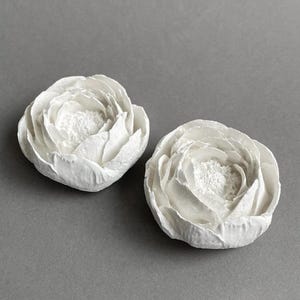Puede incluir: Dos esculturas de flores de cerámica blanca con un diseño texturizado y plisado. Las flores están cerradas y se asemejan a ranúnculos.