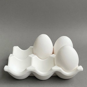 Puede incluir: Un cartón de huevos de cerámica blanca con tres huevos blancos en su interior. El cartón tiene seis compartimentos individuales para huevos.