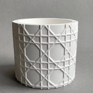 Könnte beinhalten: Ein weißer, zylindrischer Blumentopf mit geometrischem Muster. Der Topf hat eine strukturierte Oberfläche mit einem sich wiederholenden Design aus Quadraten, Achtecken und diagonalen Linien. Das Innere ist glatt und weiß.