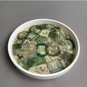 Könnte beinhalten: Eine weiße Keramik Schale gefüllt mit grün-goldenen Vintage-Aufklebern. Die Aufkleber zeigen botanische Illustrationen, Schmetterlinge und Text wie "Basil Wagon" und "Green Plant Spring".