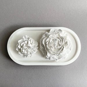 Könnte beinhalten: Ein weißes ovales Tablett mit zwei unterschiedlich großen, skulpturalen weißen Blumendekorationen. Die Blumen haben detaillierte, geschichtete Blütenblätter mit matter Oberfläche und sind auf der glatten Fläche des Tabletts platziert.