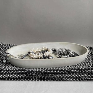 Könnte beinhalten: Eine ovale, hellgraue Betonschale mit einer Sammlung von Schmuckstücken, darunter ein weißes Perlenarmband und ein silberfarbenes Metallketten-Accessoire, die auf einer strukturierten, schwarz-weiß gewebten Stoffoberfläche liegt.