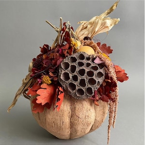 Könnte beinhalten: Herbstliche Dekoration mit einer kürbisförmigen Basis aus natürlicher Kokosnussschale. Die Anordnung ist mit getrockneten Blumen und Blättern in Rot-, Orange- und Brauntönen sowie einer einzigartigen Samenkapsel geschmückt. Die Gesamtästhetik ist rustikal und herbstlich.