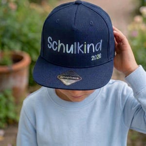 Könnte beinhalten: Eine marineblaue Snapback-Cap mit dem gestickten Schriftzug "Schulkind" in hellblauen und weißen Buchstaben sowie der darunter in Hellgrün gestickten Jahreszahl "2026". Ein rautenförmiger "Snapback"-Aufkleber befindet sich auf dem flachen Schirm.