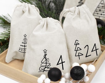 May include: Three off-white drawstring fabric pouches featuring black printed designs. One pouch shows a stylized tree, another displays a tree with the number 4, and the third features a tree with the number 24. The bags are in a wooden tray.