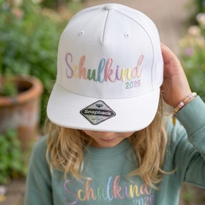 Könnte beinhalten: Eine weiße Snapback-Baseballkappe mit dem Wort "Schulkind" in Regenbogenfarben gestickt, zusammen mit dem Jahr "2026". Die Kappe hat einen flachen Schirm und einen Schnappverschluss. Die Kappe wird von einer Person gehalten.