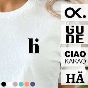 Könnte beinhalten: Weißes T-Shirt mit dem Wort "hi" in Schwarz auf der Vorderseite. Das Shirt wird an einem Model gezeigt. Das Bild zeigt auch eine Vielzahl von Grüßen in verschiedenen Sprachen, darunter "Ok", "Gude", "Ciao", "Kakao" und "H".