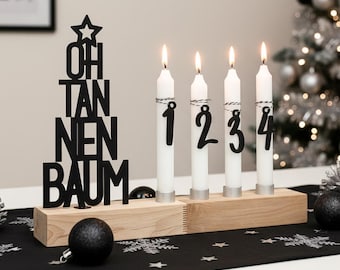 May include: A wooden advent calendar with four lit white candles, each marked with a black number from one to four. A black Christmas tree-shaped decoration with the words "OH TANNENBAUM" stands to the left. Black and silver ornaments are on a black table runner.
