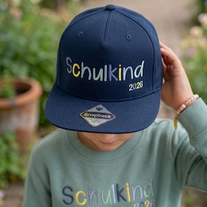 Könnte beinhalten: Marineblaue Snapback-Kappe mit dem Wort "Schulkind" in Gelb und Grün gestickt, sowie der Jahreszahl "2026". Die Kappe hat einen flachen Schirm und ein "Snapback"-Etikett.