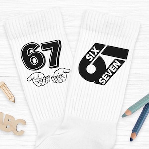 Könnte beinhalten: Paar weiße Socken mit schwarzen Grafiken. Eine Socke zeigt die Zahl "67" mit Händen, die auf die Zahlen zeigen. Die andere Socke zeigt "SIX SEVEN" in einem stilisierten Design. Die Socken liegen auf einer weißen Holzoberfläche.