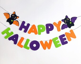 May include: A felt Halloween banner with the words "Happy Halloween" in orange, green, and purple letters. Two black bats with orange wings and pink hearts are on either end of the banner.