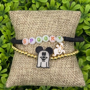 Op de afbeelding: Twee Halloween-armbanden. De bovenste armband is zwart met het woord "SPOOKY" gespeld in kleurrijke letterkralen. De onderste armband is goudkleurig met een Mickey Mouse spookbedel en een spookbedel.