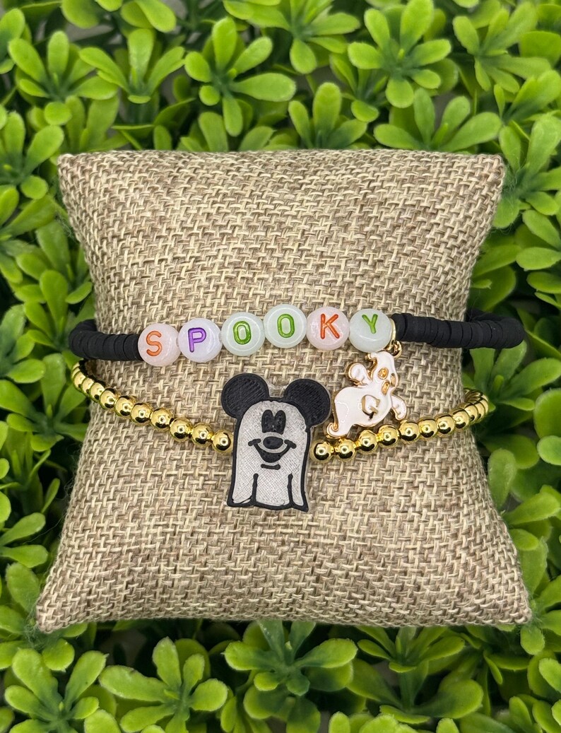 Op de afbeelding: Twee Halloween-armbanden. De bovenste armband is zwart met het woord "SPOOKY" gespeld in kleurrijke letterkralen. De onderste armband is goudkleurig met een Mickey Mouse spookbedel en een spookbedel.