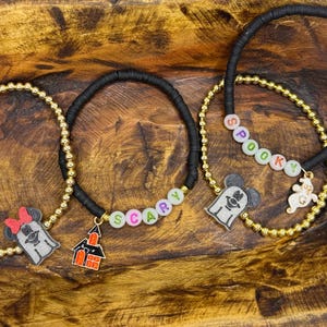 Op de afbeelding: Drie Halloween-armbanden. Een goudkleurige kralenarmband met een Minnie Mouse spookbedel en een zwarte armband met het woord "SCARY" in kleurrijke kralen. Een andere goudkleurige kralenarmband met een spookbedel en een zwarte armband met het woord "SPOOKY".