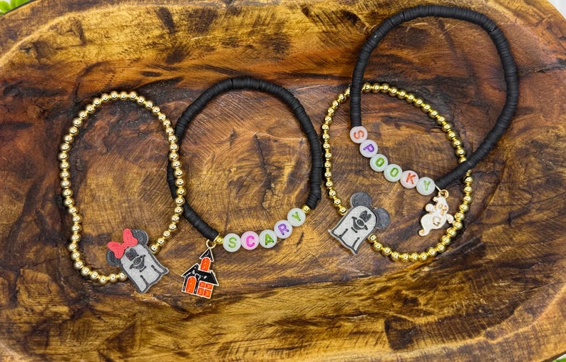Op de afbeelding: Drie Halloween-armbanden. Een goudkleurige kralenarmband met een Minnie Mouse spookbedel en een zwarte armband met het woord "SCARY" in kleurrijke kralen. Een andere goudkleurige kralenarmband met een spookbedel en een zwarte armband met het woord "SPOOKY".