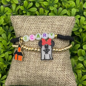 Op de afbeelding: Twee Halloween-armbanden. E&eacute;n armband heeft het woord "SCARY" in letterkralen. De andere armband heeft een gouden kralenketting met een spook- en spookhuisbedel. De armbanden worden op een jute kussen getoond.