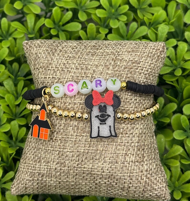 Op de afbeelding: Twee Halloween-armbanden. E&eacute;n armband heeft het woord "SCARY" in letterkralen. De andere armband heeft een gouden kralenketting met een spook- en spookhuisbedel. De armbanden worden op een jute kussen getoond.
