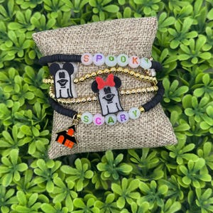 Op de afbeelding: Een stapel Halloween-armbanden. E&eacute;n armband spelt "Spooky" in kleurrijke kralen. Een andere armband spelt "Scary". Elke armband heeft een zwart Mickey Mouse-silhouet en een kleine oranje spookhuis-bedel. De armbanden liggen op een jute oppervlak.
