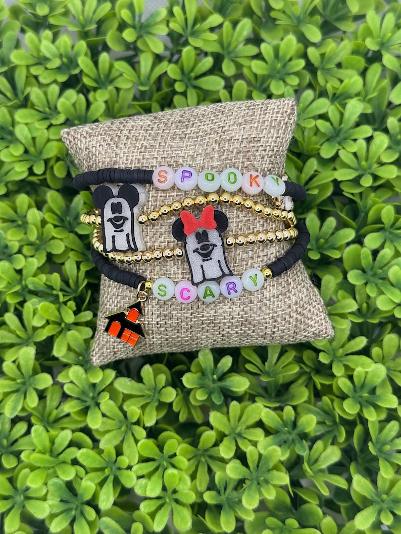 Op de afbeelding: Een stapel Halloween-armbanden. E&eacute;n armband spelt "Spooky" in kleurrijke kralen. Een andere armband spelt "Scary". Elke armband heeft een zwart Mickey Mouse-silhouet en een kleine oranje spookhuis-bedel. De armbanden liggen op een jute oppervlak.