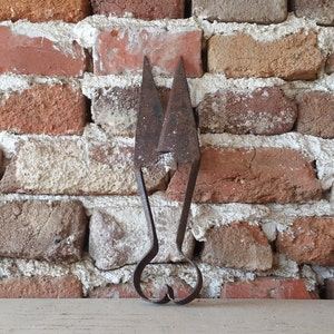 May include: A pair of antique rusty metal shears with a heart-shaped handle. The shears are against a brick wall background.