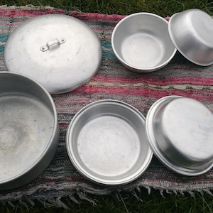 Könnte beinhalten: Ein Satz aus fünf silbernen Aluminiumschalen und Deckeln. Die Schalen sind ineinander verschachtelt auf einem bunten gestreiften Teppich.