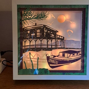 Peut inclure: Une boîte à ombre encadrée de blanc avec une bordure à motifs verts et bruns. La boîte contient une scène découpée en papier d'une maison sur pilotis au-dessus de l'eau, un bateau avec le mot "DINBOU" dessus, un soleil et des oiseaux. La scène est éclairée par des lumières blanches chaudes.