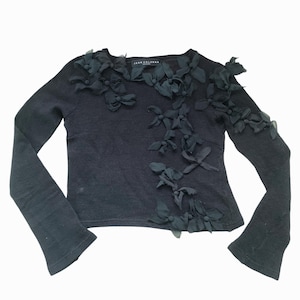 A black long-sleeved sweater with a crew neck and black fabric flowers on the front. The sweater has the words 'JEAN COLONNA' on the label.
