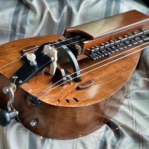 May include: A wooden hurdy-gurdy instrument with a curved body and a keyboard. The instrument has a dark brown finish with visible wood grain. It features strings, a crank, and a series of keys. The instrument is designed for playing music.