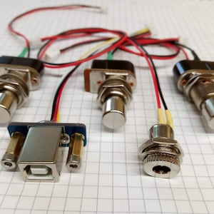 Puede incluir: Cinco interruptores de botón pulsador plateados con cables rojos, negros y amarillos. Los interruptores están conectados a una pequeña placa de circuito impreso con un conector USB.