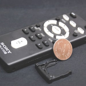 May include: A black remote control with a silver button labelled "ENTER" and a white button labelled "VOL+". The remote has a silver battery labelled "CR2025 SONY" and the text "SONY RM-X153" printed on the bottom.