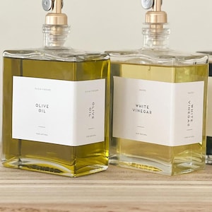 May include: Two clear glass bottles with gold tops and labels. The bottle on the left is filled with olive oil and the label reads "Olive Oil". The bottle on the right is filled with white vinegar and the label reads "White Vinegar".