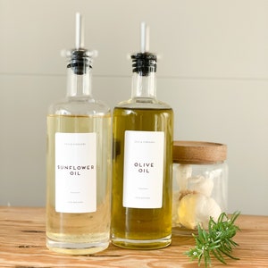 K&ouml;nnte beinhalten: Zwei Glasflaschen mit Ausgie&szlig;ern, eine gef&uuml;llt mit Sonnenblumen&ouml;l und die andere mit Oliven&ouml;l. Beide Flaschen tragen Etiketten mit der Aufschrift "Oils & Vinegars" und der &Ouml;lsorte. Die Flaschen stehen auf einer Holzoberfl&auml;che neben einem Glasgef&auml;&szlig; mit Knoblauchzehen und einem Rosmarinzweig.