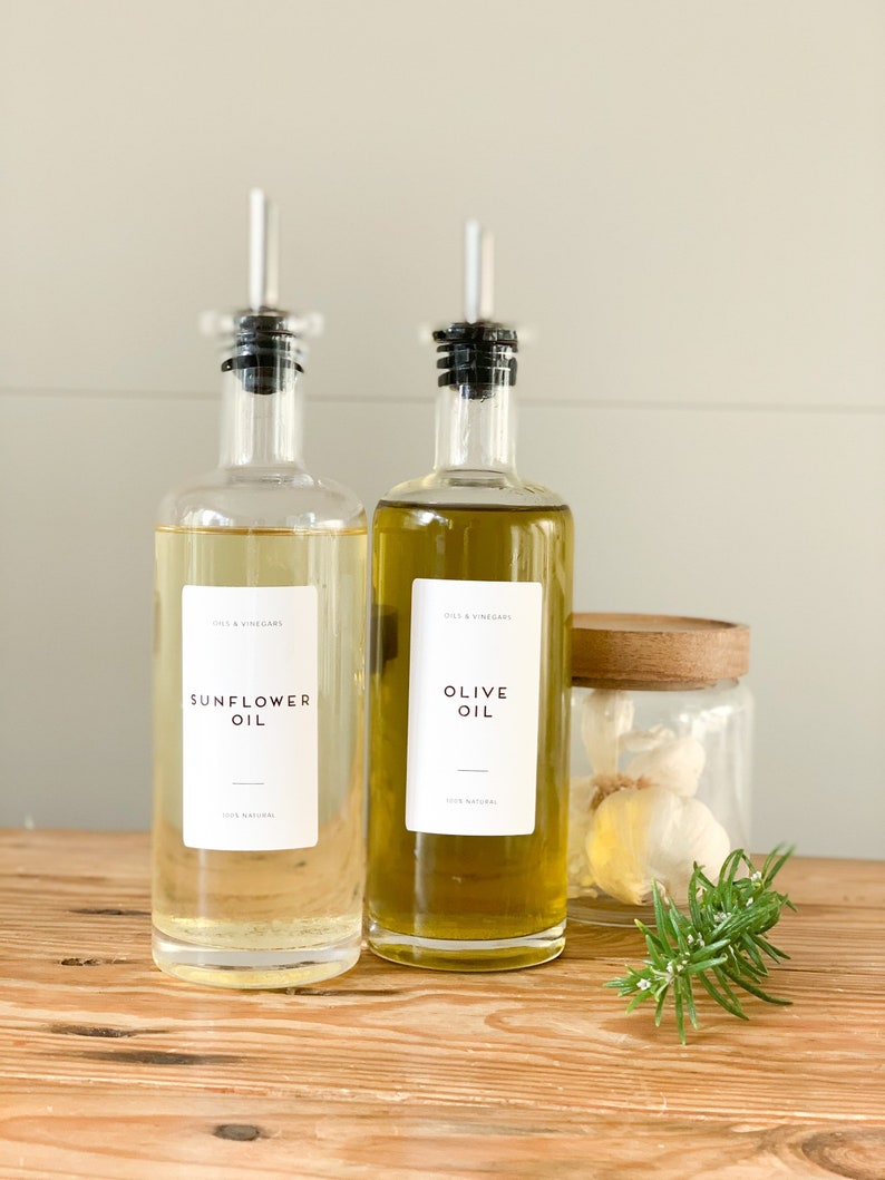 K&ouml;nnte beinhalten: Zwei Glasflaschen mit Ausgie&szlig;ern, eine gef&uuml;llt mit Sonnenblumen&ouml;l und die andere mit Oliven&ouml;l. Beide Flaschen tragen Etiketten mit der Aufschrift "Oils & Vinegars" und der &Ouml;lsorte. Die Flaschen stehen auf einer Holzoberfl&auml;che neben einem Glasgef&auml;&szlig; mit Knoblauchzehen und einem Rosmarinzweig.