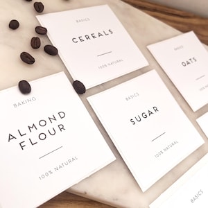 May include: White rectangular pantry labels with black text. Labels include "CEREALS", "ALMOND FLOUR", "SUGAR", and "OATS". The labels also include the text "BASICS" and "100% NATURAL". Several coffee beans are scattered around the labels.