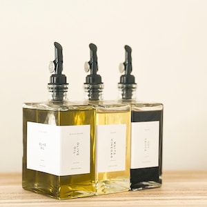 May include: Three glass bottles with black pour spouts and white labels. The bottles contain olive oil, white vinegar, and soy sauce. The bottles are sitting on a wooden surface.