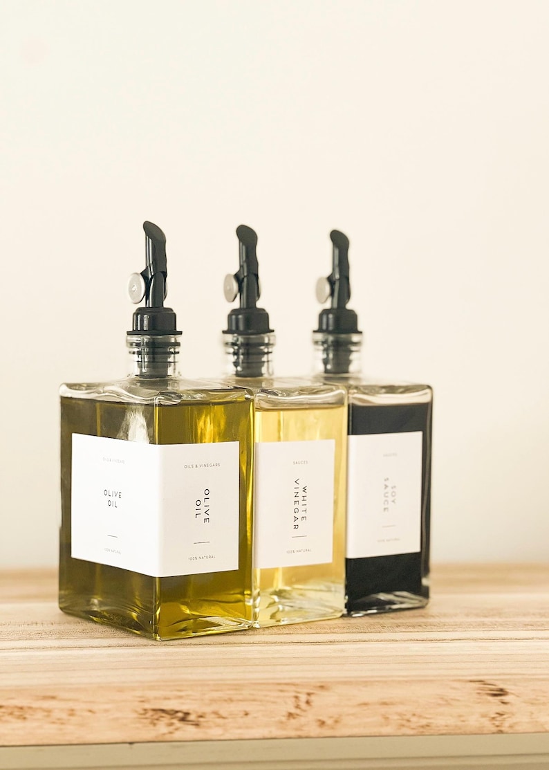 May include: Three glass bottles with black pour spouts and white labels. The bottles contain olive oil, white vinegar, and soy sauce. The bottles are sitting on a wooden surface.