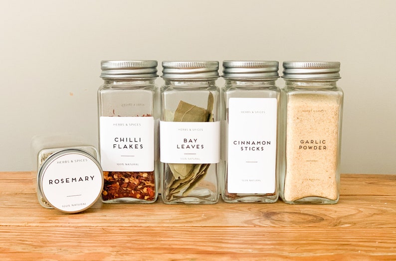 K&ouml;nnte beinhalten: F&uuml;nf Glasbeh&auml;lter mit silbernen Deckeln, die verschiedene Kr&auml;uter und Gew&uuml;rze enthalten. Die Beh&auml;lter sind mit wei&szlig;en Etiketten beschriftet, auf denen "Rosmarin", "Chili-Flocken", "Lorbeerbl&auml;tter", "Zimtstangen" und "Knoblauchpulver" steht.