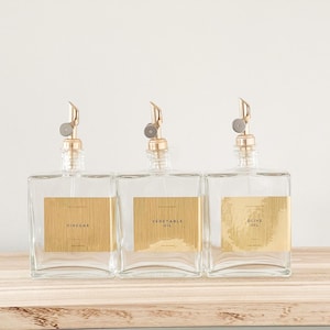 May include: Three clear glass bottles with gold spouts and labels. The labels read "Vinegar", "Vegetable Oil", and "Olive Oil".