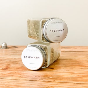 May include: Two clear, square spice jars stacked on a wooden surface. The top jar is labeled "Oregano" and the bottom jar is labeled "Rosemary". Both jars have silver lids with the text "Herbs & Spices" and "100% Natural".