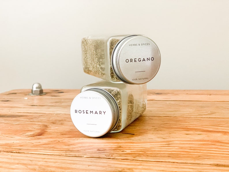 May include: Two clear, square spice jars stacked on a wooden surface. The top jar is labeled "Oregano" and the bottom jar is labeled "Rosemary". Both jars have silver lids with the text "Herbs & Spices" and "100% Natural".