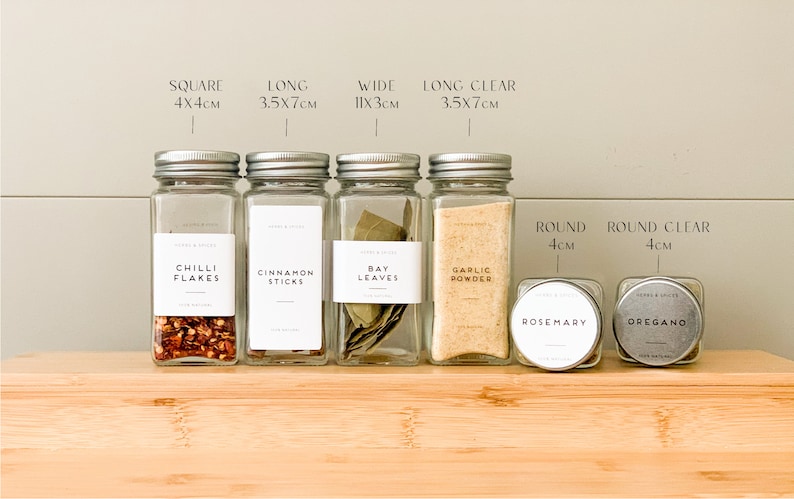 May include: A selection of glass spice jars with silver lids, labelled with the names of herbs and spices. The jars are arranged on a wooden surface, with measurements in centimetres displayed above.