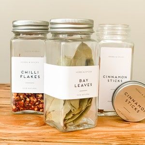 May include: A selection of glass spice jars and a small tin on a wooden surface. The jars contain chilli flakes, bay leaves, and cinnamon sticks. Each jar has a white label with the text "HERBS & SPICES" and the spice name. The text "100% NATURAL" is also visible.