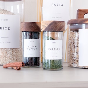 May include: A set of glass jars with wooden lids labelled with the words "Rice", "Black Pepper", "Parsley", and "Oats". The jars are filled with various pantry staples, including rice, black peppercorns, dried parsley, and oats. The labels are all white with black text and a thin black line underneath the text. The jars are arranged on a white countertop.