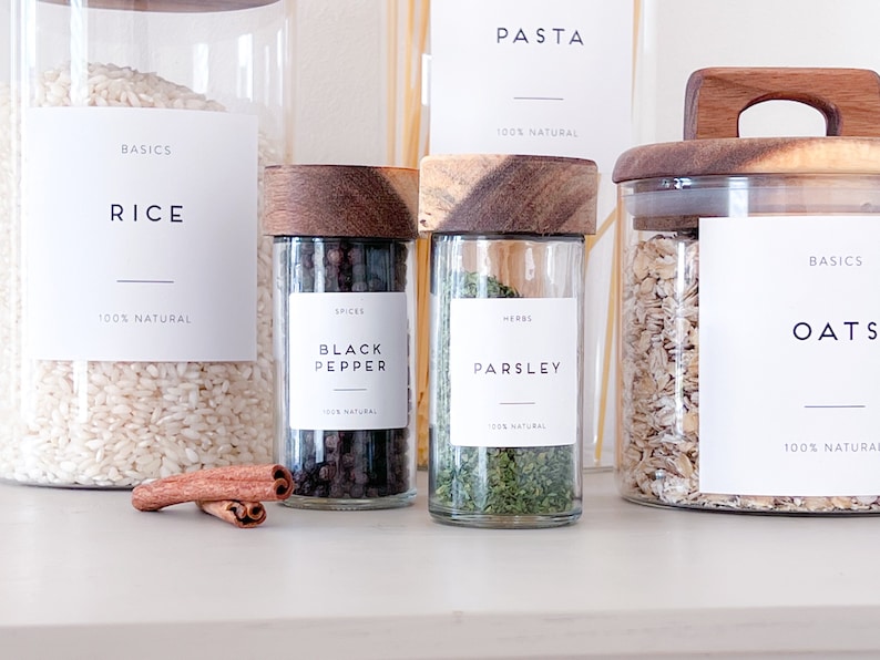 May include: A set of glass jars with wooden lids labelled with the words "Rice", "Black Pepper", "Parsley", and "Oats". The jars are filled with various pantry staples, including rice, black peppercorns, dried parsley, and oats. The labels are all white with black text and a thin black line underneath the text. The jars are arranged on a white countertop.