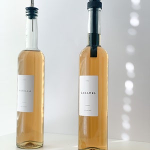May include: Two glass bottles with brown liquid and black pour spouts. The bottles have white labels with the words "Vanilla" and "Caramel" printed on them. The bottles are sitting on a white surface with coffee beans scattered in front of them.